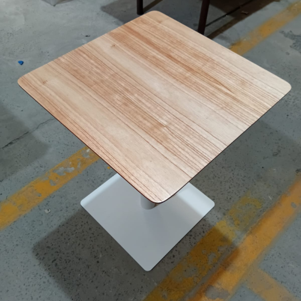 Mesa cuadrada con tapa de madera Paraíso veteada y base central de caño metálico blanco sobre un piso de fábrica.