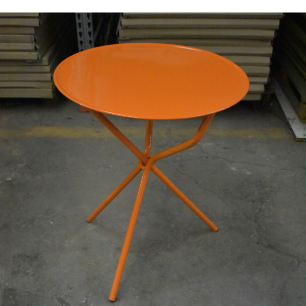 Mesa redonda de metal color naranja, modelo Imperial, con patas de caño cruzadas sobre un piso de cemento de fábrica.