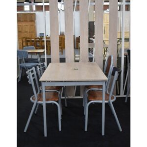 Juego de comedor con mesa cuadrada de madera clara y cuatro sillas de caño gris con asientos marrones en una exposición.